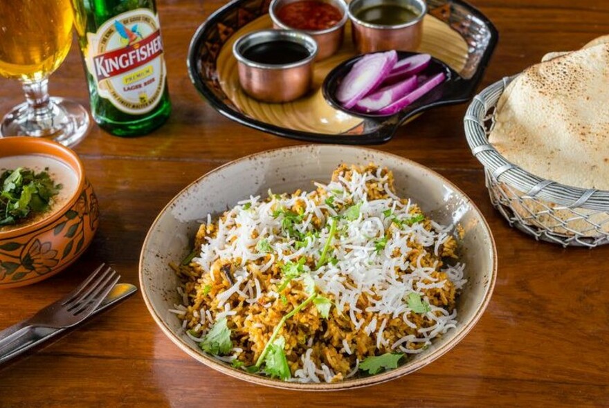 A bowl of curry and rice with Kingfisher beer, bread and condiments.