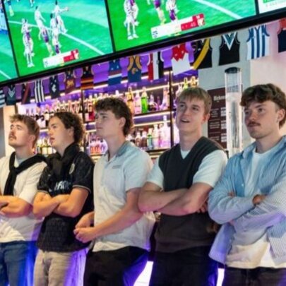 Five young men leaning against a sports bar with their arms crossed, multiple television screens behind them showing football.