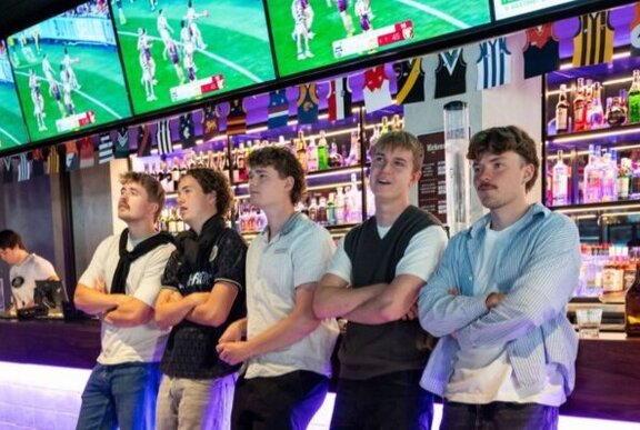 Five young men leaning against a sports bar with their arms crossed, multiple television screens behind them showing football.