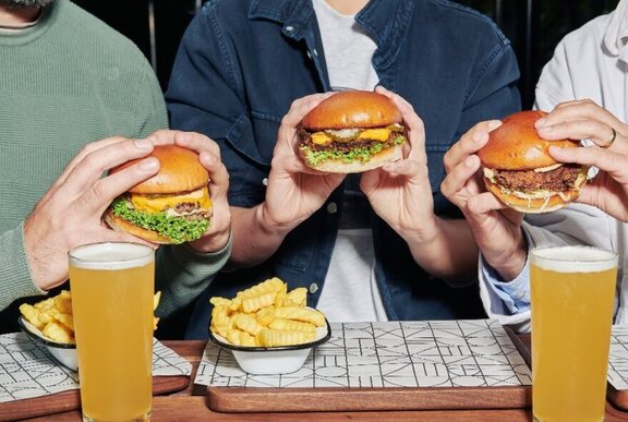 Three pairs of hands holding up three juicy cheeseburgers, with a bowl of crinkle cut chips and two glasses of beer on a table.