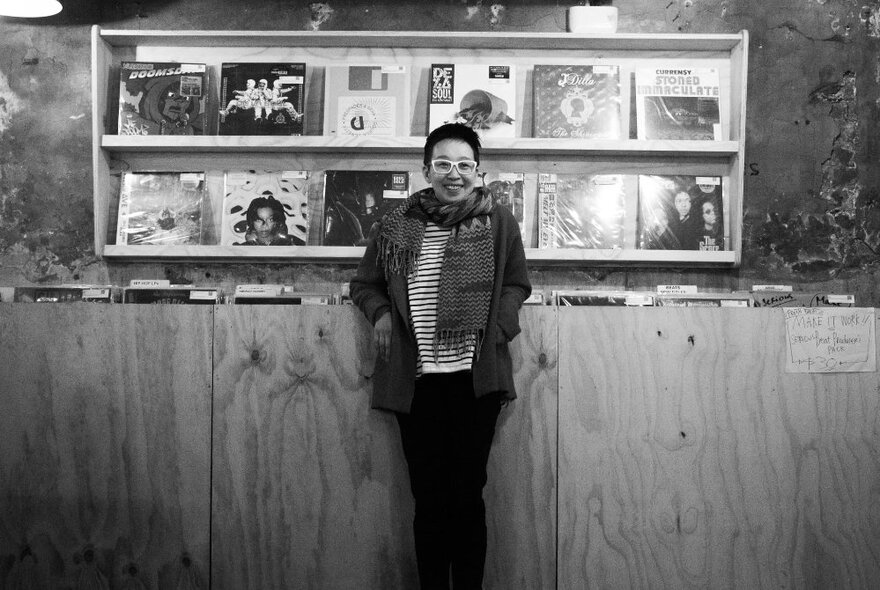 A smiling person wearing glasses and a scarf, leaning against a wooden counter in a record store; black and white image.