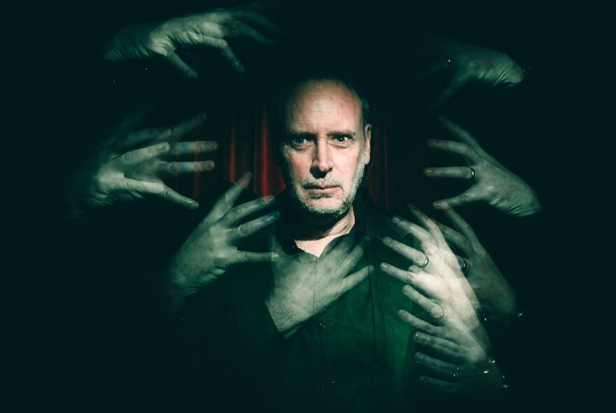 A spooky image of a man in front of a red curtain, with multiple blurred hands surrounding his face.