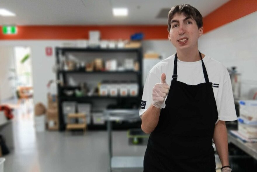A hospitality worker wearing an apron, smiling and standing inside a kitchen.