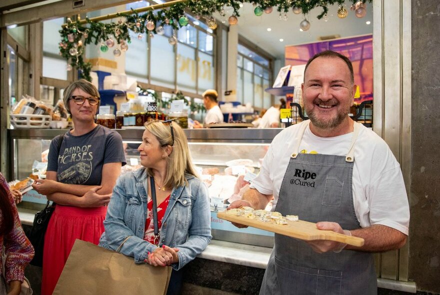 A merchant at the Queen Victoria Market holding a wooden board with cheese samples, while tour participants lean against the counter of his market stall.