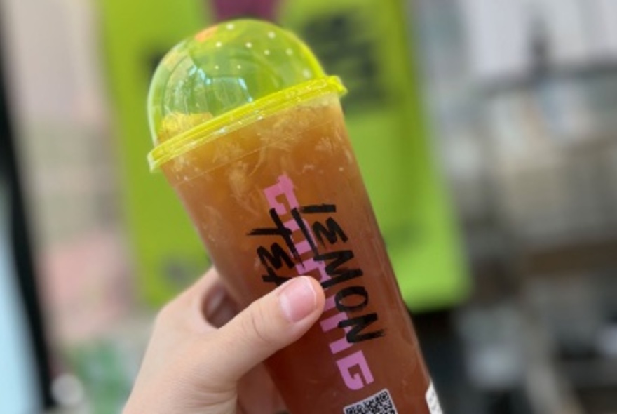 Hand holding a plastic takeaway cup of iced lemon tea with a domed lid on the top of the cup.