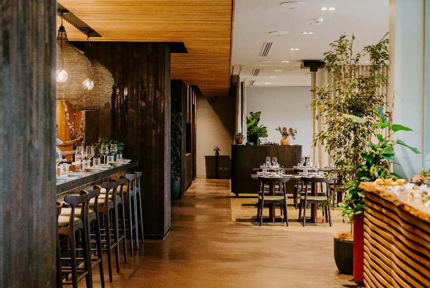 Interior of restaurant showing tables set for service in the background, greenery, and in the foreground a area to sit at a high bar.