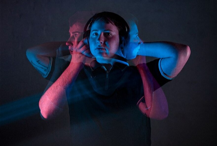 A blurred image of a person holding their hands to their ears against a dark background.