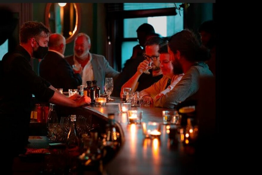 Curved wooden bar, decorated with small tealight candles, with patrons standing at the drinking and a bartender serving drinks.