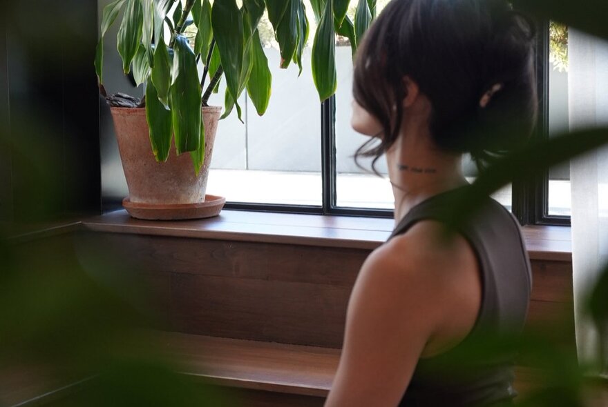 Side view of a person who is sitting quietly near a window, with a potted plant resting on the window sill.