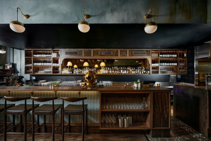 Interior of restaurant showing a bar counter,  high stools, bottles on shelves, and three   lightshades hanging overhead.