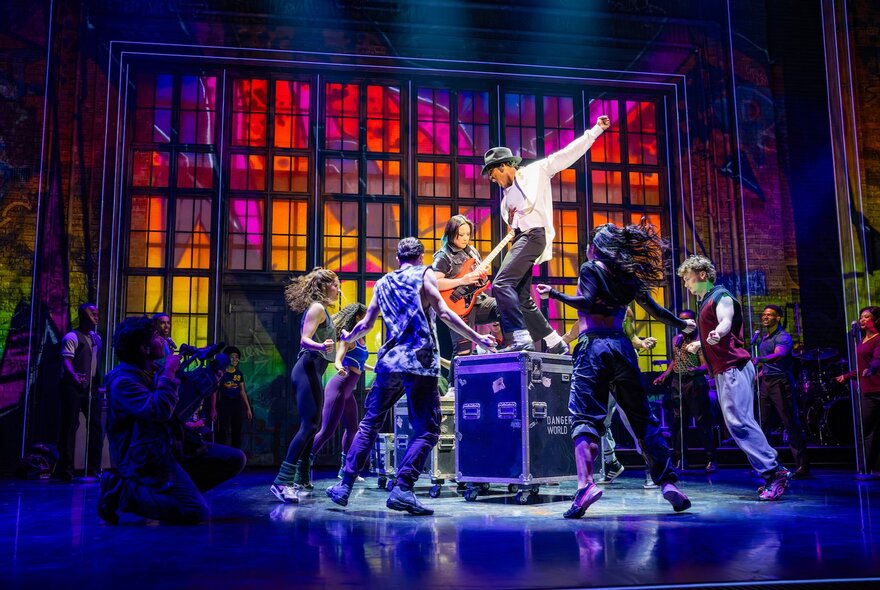 A dancer on stage in a white jacket and black pants, in the role of Michael Jackson, with musicians and dancers alongside, in the stage production of MJ the Musical.