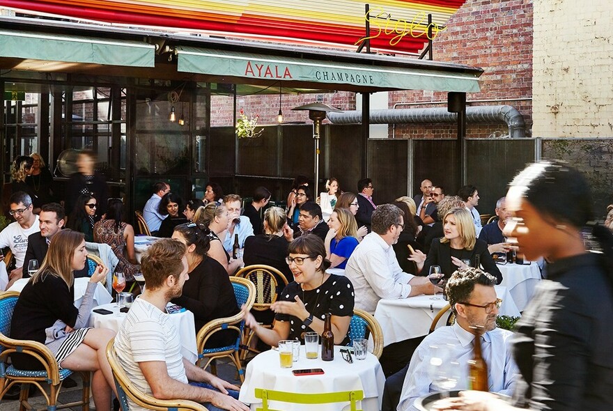 Siglo outdoor area crowded with people eating and talking at tables with wicker chairs.