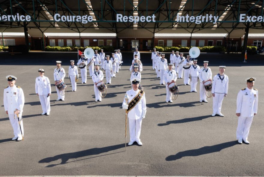 Member of the Royal Australian Navy Band, dressed in white, standing outdoors in lines.