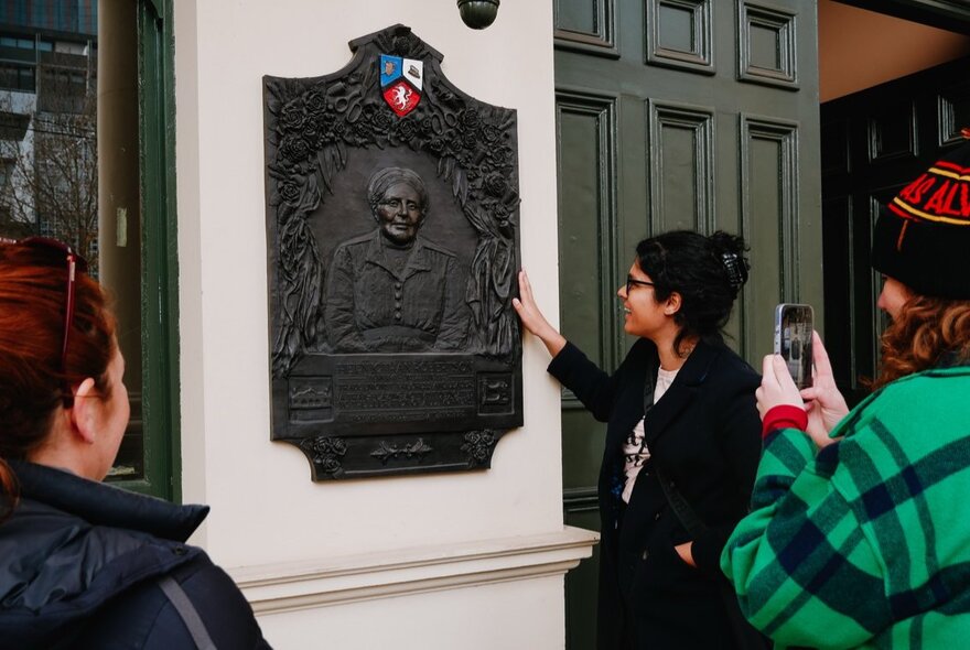 A tour guide talking and pointing to a plaque on a building's wall, with tour group participants listening and taking photos. 