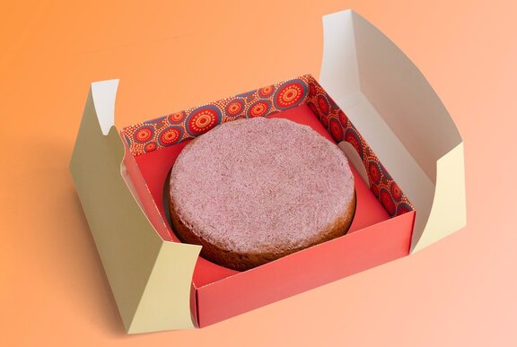 A white gateau cake dusted with pink icing, in a natural red-coloured box with Indigenous designs on the sides.