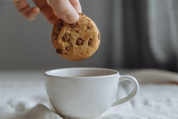 A hand holding a choc-chip biscuit above a cup of hot tea, as if about to dip it in.