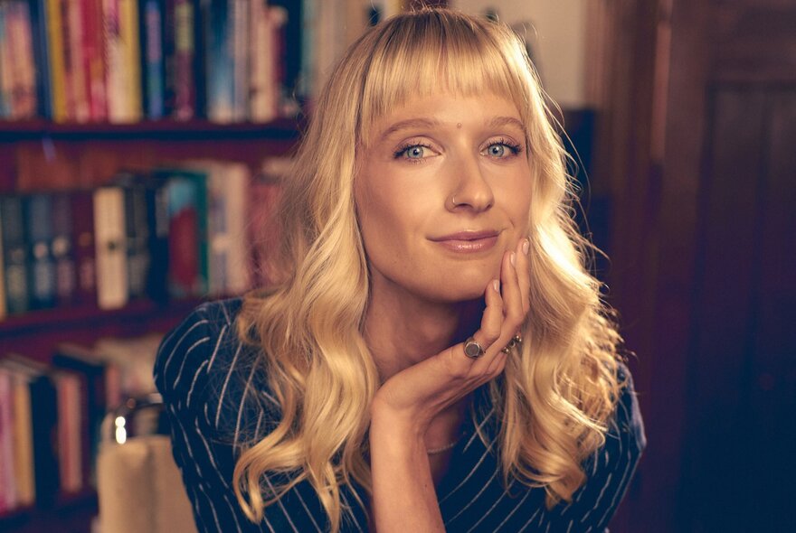 Blonde woman with long hair resting her head on her hand, bookshelves in the background.