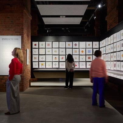 People looking at a gallery wall of small white square embroidered handkerchiefs. 