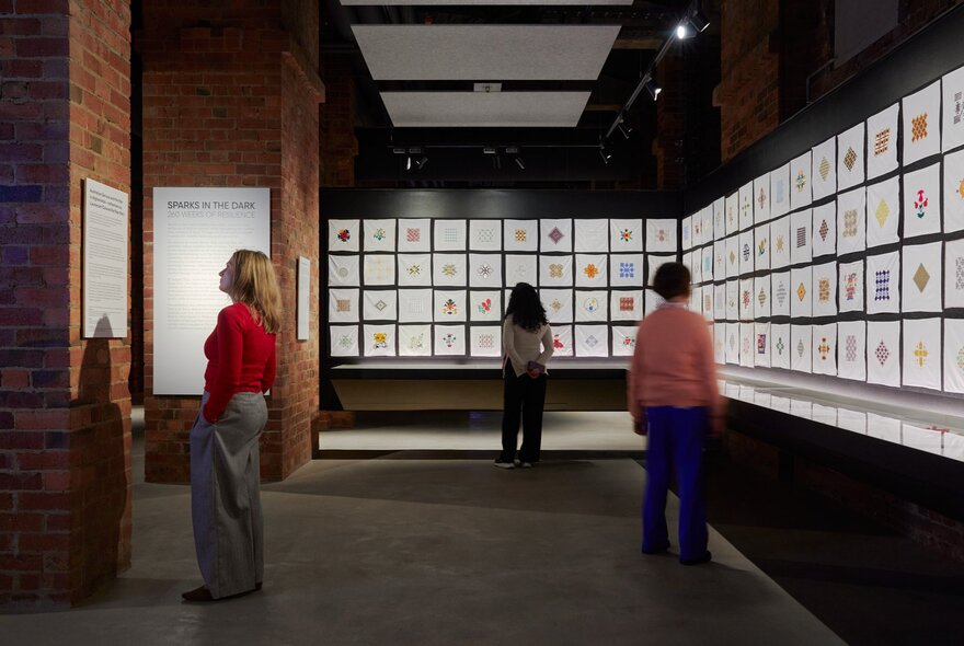 People looking at a gallery wall of small white square embroidered handkerchiefs. 
