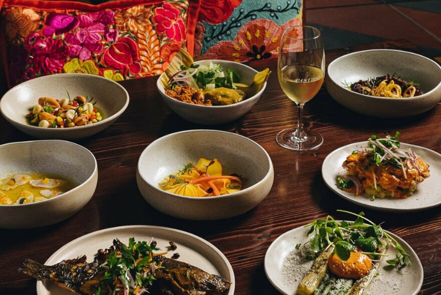 Table set with many dishes including salads, meats, a whole fish, dips and a glass of wine.