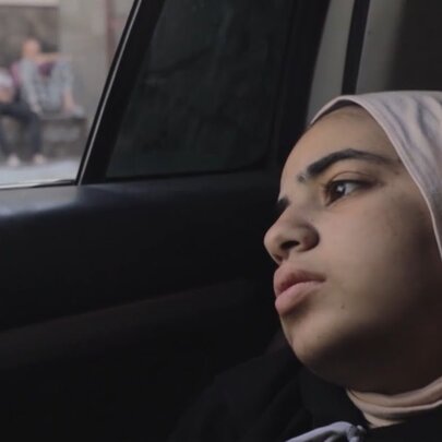 Film still of a young girl wearing a headscarf, leaning her head against the door in a car.