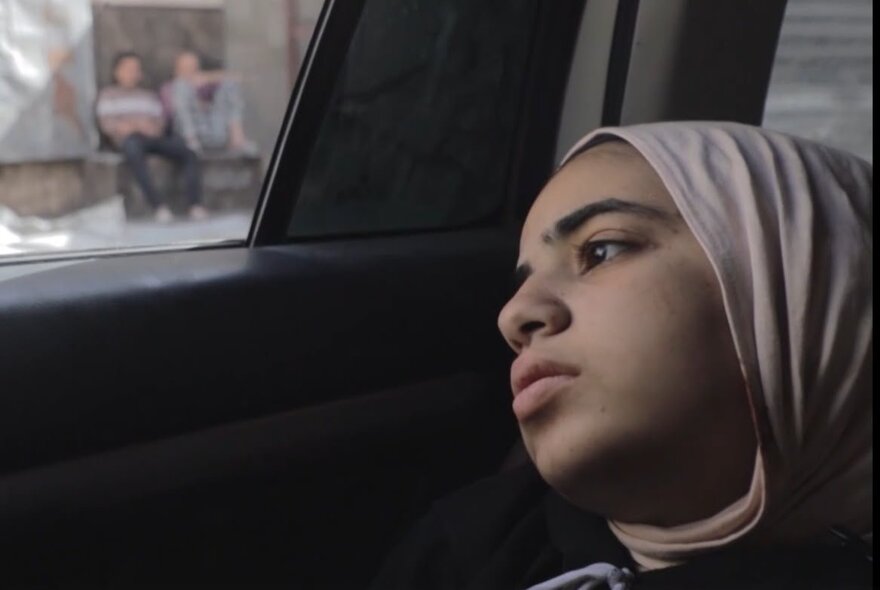 Film still of a young girl wearing a headscarf, leaning her head against the door in a car.