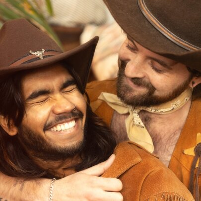 Two people dressed as cowboys wearing cowboy hats sharing a warm, laughing embrace.