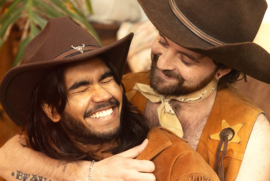 Two people dressed as cowboys wearing cowboy hats sharing a warm, laughing embrace.