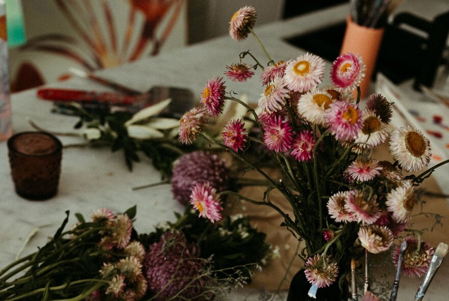 A close-up of a floral arrangement workshop table with a vase of pink and white strawflowers, with art materials in the background.