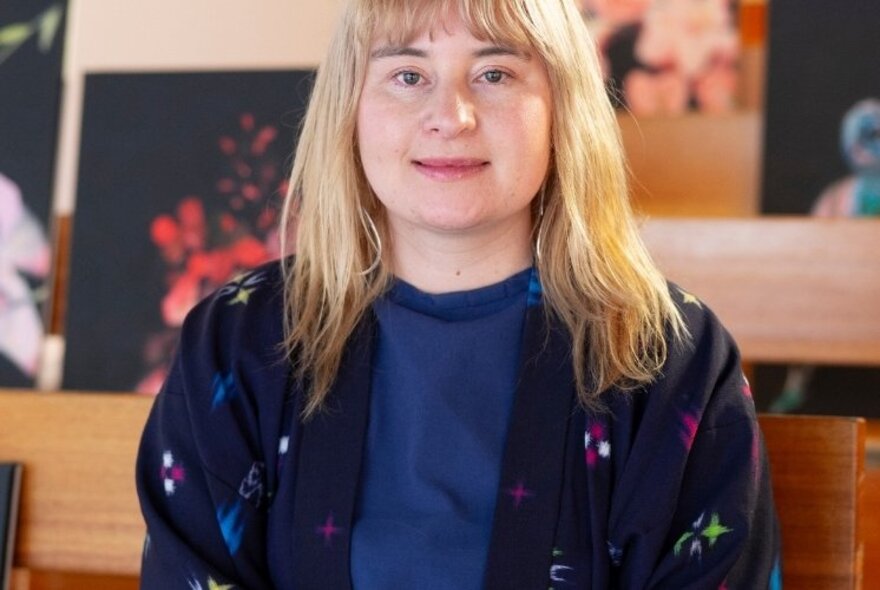 Young person with shoulder length straight blond hair, wearing a patterned jacket, seated on a wooden bench with colourful artworks in the background.