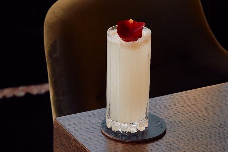 A tall white cocktail with a deep red rose petal garnish on a dark wood table. 