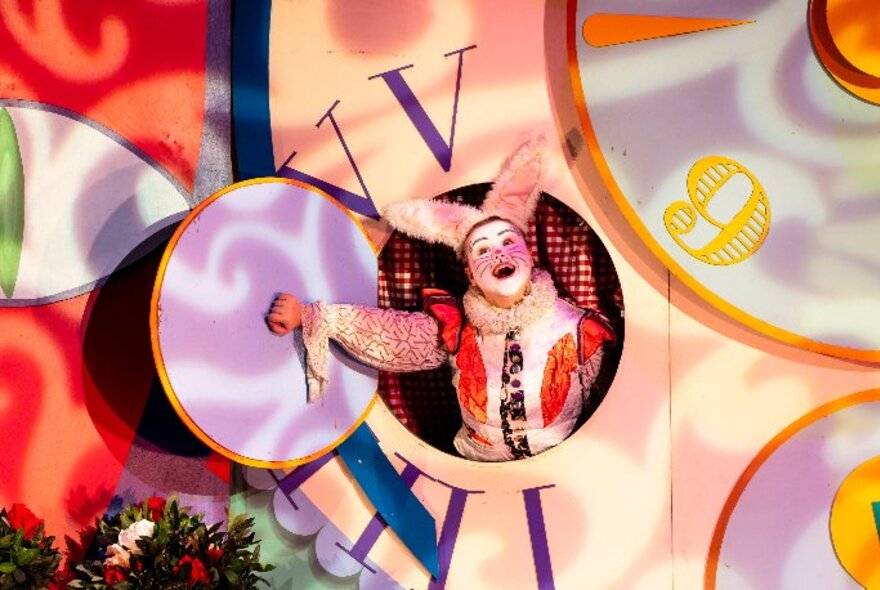 An actor in the role of the White Rabbit performing in the family theatre show of Alice in Wonderland, poking their head out of a giant clock prop on stage.