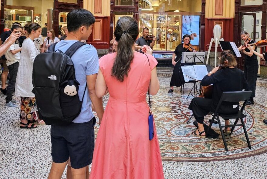 A young couple watching a string quartet perform inside a mosaic-tiled arcade.