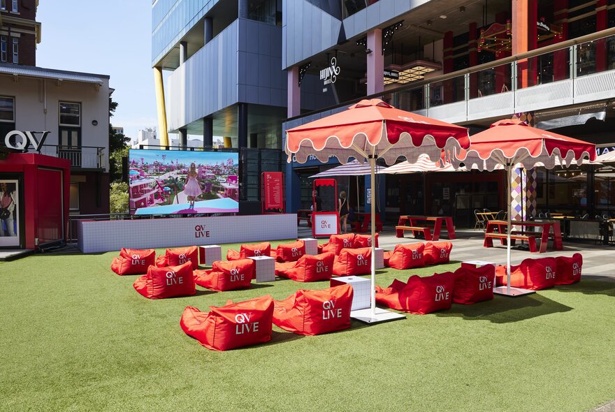 Red bean bag chairs under red fringed umbrellas on a surface of fake grass with city buildings all around. 
