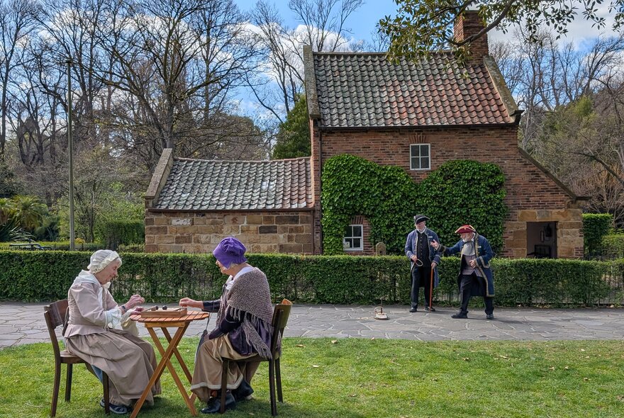 People dressed up in 1700s costumes outside Cook's Cottage in Fitzroy Gardens, the women taking tea and the two men playing quoits. 