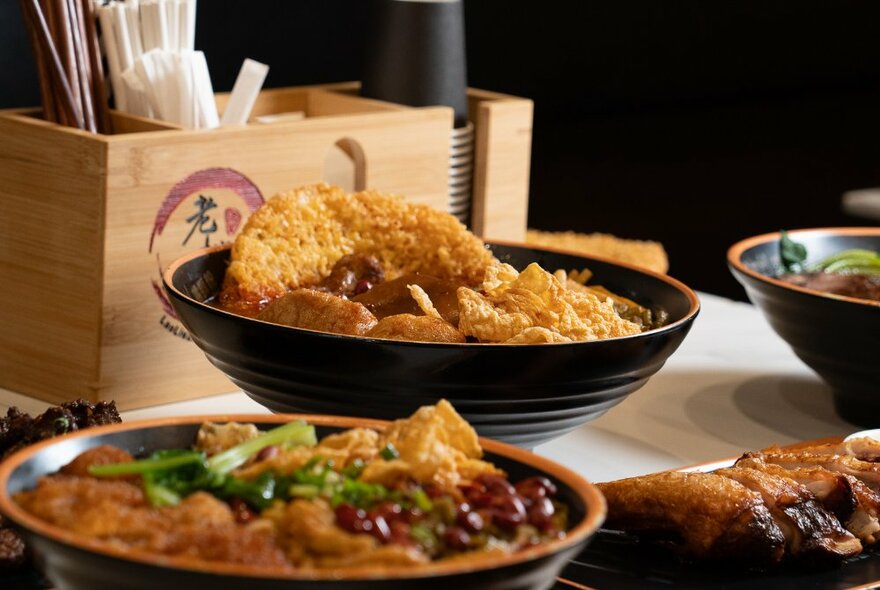 Two bowls of steaming noodle dishes on a table, with a box of dining accessories in the background.