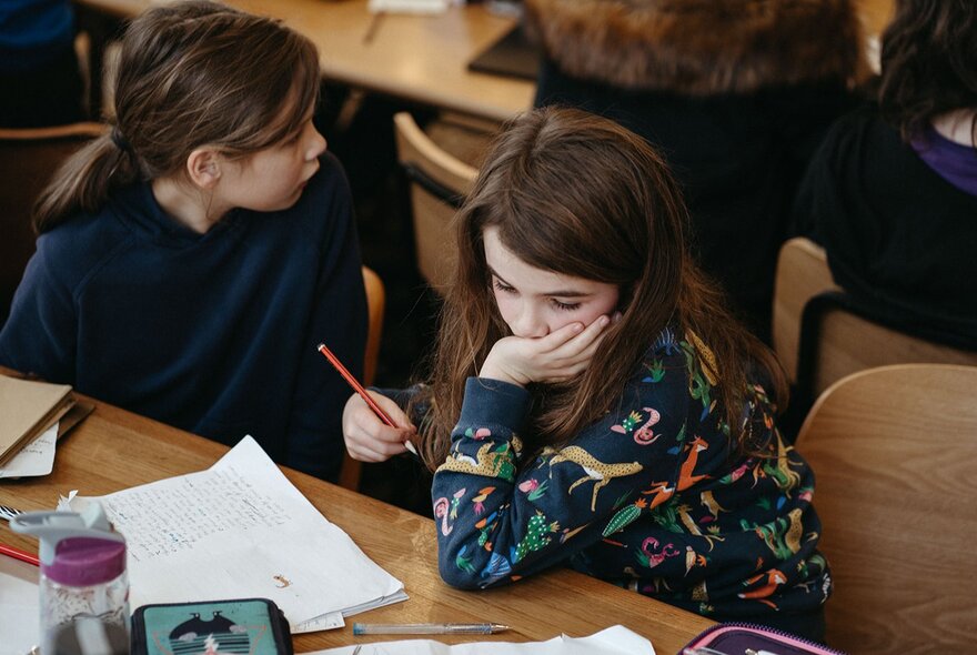 Children seated at a table, one with her head in her hands, holding a pen with paper.
