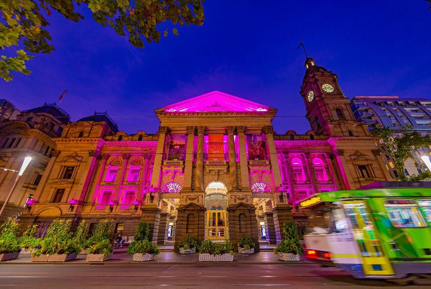 Melbourne Town Hall at night with pink lights illuminating the front facade and a tram passing by.