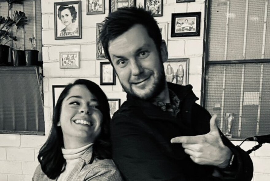 A man and woman standing back to back and smiling, in a room with pictures on the wall behind them, black and white image.