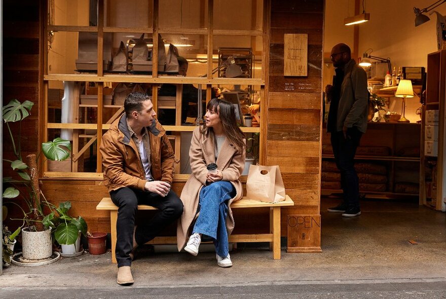 A man and woman sitting on a bench seat outside a timber bakery. 