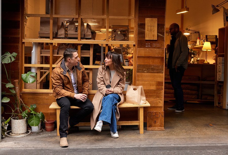 A man and woman sitting on a bench seat outside a timber bakery. 