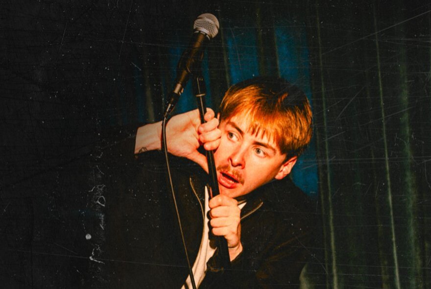 Comedian, Blake Pavey, crouching behind a microphone stand on stage as though hiding.