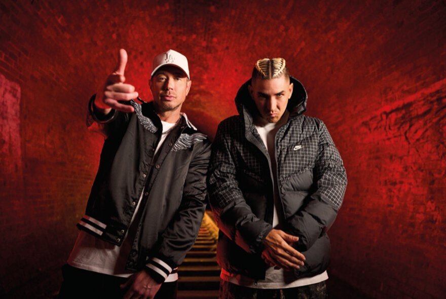 Hip hop duo Bliss N Eso, both wearing dark jackets, one of them with their arm stretched out in front of them pointing at the viewer, posed against a red background.