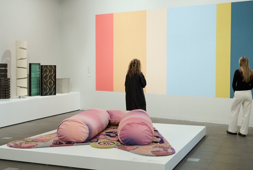 Two people are looking at a colourful striped painting on a wall in a gallery. There is a large pink, soft blobby sculpture behind them.