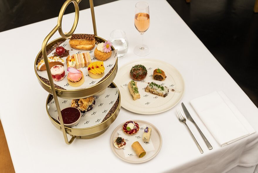 A gold two-tiered cake stand alongside two small white plates, all filled with a selection of small delicacies.