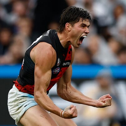 An Essendon AFL player cheering himself and the crowd on with pumped fists, on the footy field.