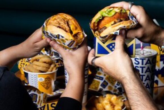 Two people's hands holding burgers, with buckets of chips in the background.