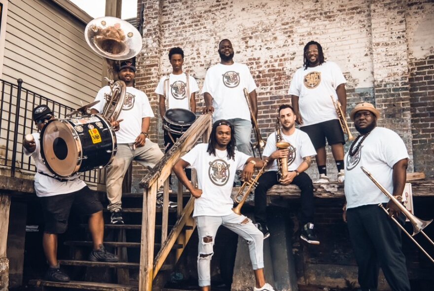 The members of the TBC Brass Band wearing matching white t-shirts with a band slogan printed on the font, posed as a group standing on low steps in front of a faded brick wall and holding their instruments.