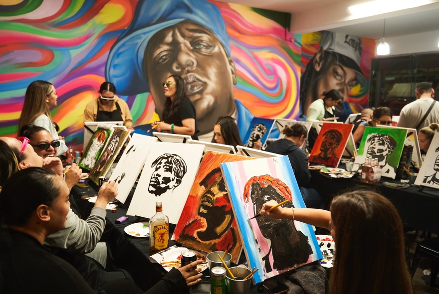 Several rows of people painting on canvases in a room with large, wall murals.