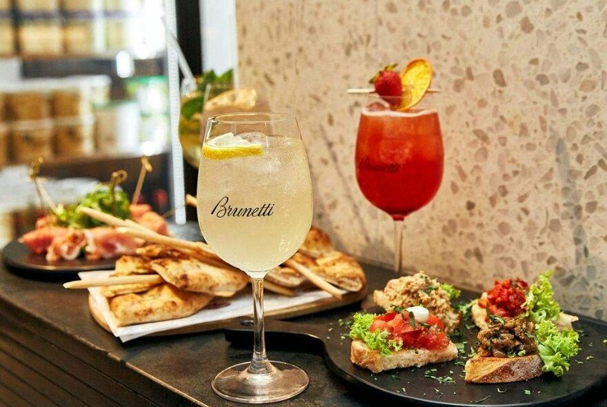 Two cocktails on a bench with plates of food beside them